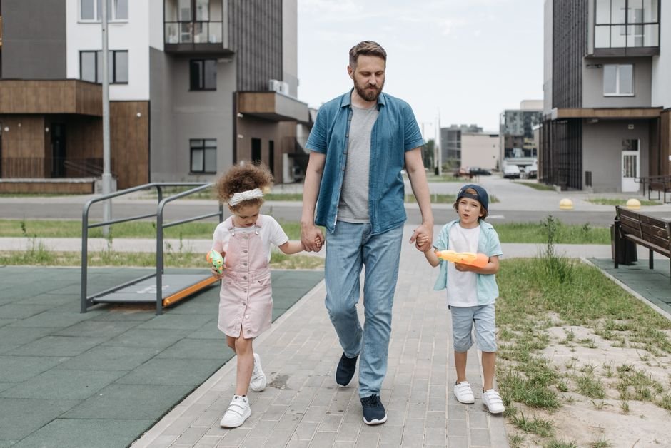 Tėtis su vaikais eina urbanizuota gyvenamąja zona link modernios žaidimų aikštelės