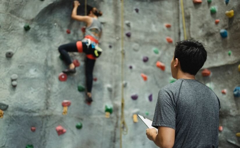 Treneris stebi mergaitę, kopiančią boulderingo sienele, stiprindama vaikų augimą
